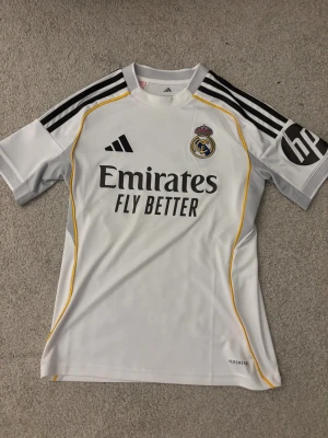 Real Madrid vit matchtröja Adidas - Säljer en officiell Real Madrid fotbollströja från Adidas i storlek 152. Tröjan är vit med svarta och gula detaljer, har korta ärmar och klubbmärket broderat på bröstet. Materialet är lätt och andas, perfekt för träning eller match. Snygga svarta ränder på axlarna och HP-logga på ärmen.