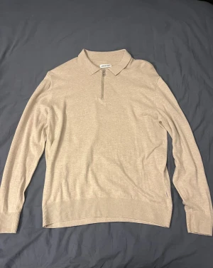 Beige stickad half zip tröja från Jack & Jones - Snygg beige tröja från Jack & Jones i storlek M. Tröjan har krage och en halv dragkedja framtill, vilket ger en clean och stilren look. Tillverkad i mjukt material med ribbade muddar vid ärmslut och nederkant. Perfekt för lager-på-lager under höst och vår.
