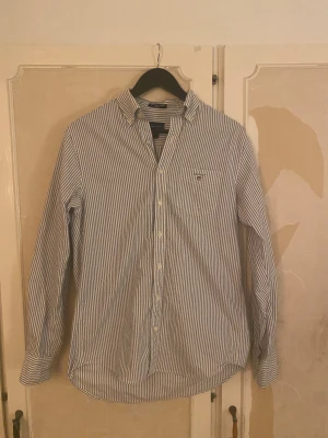 Randig Oxfordskjorta från GANT - Snygg Oxfordskjorta från GANT i storlek S, med klassiskt blåvitt randigt mönster och broderad logga på bröstfickan. Skjortan har lång ärm, knappar framtill och är tillverkad i mjuk bomull. Regular fit för en chill och stilren look.
