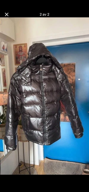 Svart pufferjacka med huva - Säljer en svart pufferjacka med glansig finish och stor huva. Jackan har dragkedja framtill, fickor med dragkedja och en extra ficka med knapp på ärmen. Passar både man och kvinna. Storlek M. Aldrig använd 