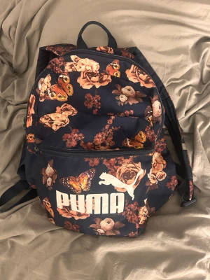 Blommig ryggsäck från Puma - Snygg ryggsäck från Puma med mörkblå botten och stora beige och bruna blommor samt fjärilar som mönster. Väskan har justerbara axelremmar, ett stort huvudfack och en framficka med dragkedja. Perfekt för skolan eller häng med kompisar.