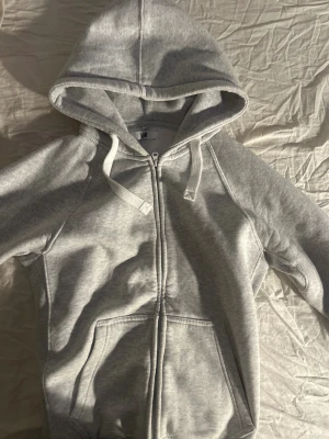 Grå hoodie från Russedress med dragkedja - Säljer en ljusgrå hoodie från Russedress i storlek M. Tröjan har huva med snörning, praktiska fickor framtill och hel dragkedja. Mjuk och skön bomullsblandning, perfekt för chill dagar. Diskret logga på ärmen och ribbade muddar vid ärmslut och nederkant.