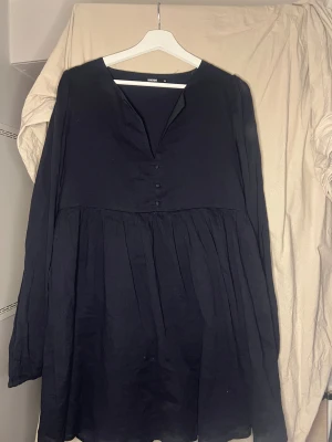 Svart långärmad klänning från Bikbok - Svart klänning från Bikbok i storlek M med lång ärm och v-ringad hals. Klänningen har tre dekorativa knappar framtill och ett lätt rynkat fall som ger en skön volym. Perfekt för dig som gillar enkel och stilren design.
