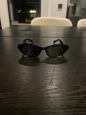 Svarta cat eye solglasögon Miu Miu - Snygga svarta solglasögon från Miu Miu med cat eye-form och guldiga logodetaljer på skalmarna. Glasögonen har en stilren och trendig look som verkligen sticker ut. Perfekta för dig som vill ha en statement-accessoar.