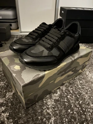 Valentino Rockrunner sneakers - Svarta Valentino Garavani Rockrunner sneakers i storlek 43 med coolt kamouflagemönster och snygga nitar på hälen. Skorna har snörning, rund tå och platt sula. Materialmix av skinn och textil ger en lyxig känsla och street vibe. Perfekt för dig som gillar exklusiva sneakers med edge. Endast använda 2gånger.                                                      Finns inga skador eller något liknande                 Vid funderingar är det bara att höra av sig :)