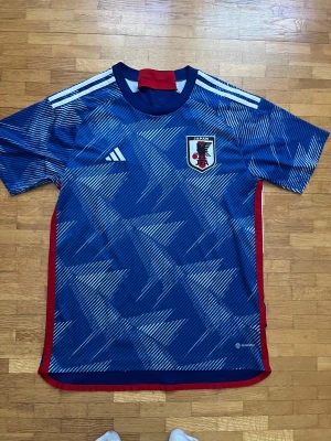 Japan blå fotbollströja Adidas L - Säljer en officiell Japan fotbollströja från Adidas i storlek L. Tröjan är blå med vita och ljusblå diagonala ränder, har korta ärmar och röda detaljer vid krage och sidor. Materialet är lätt och andas, perfekt för match eller träning. Japan-emblem på bröstet.