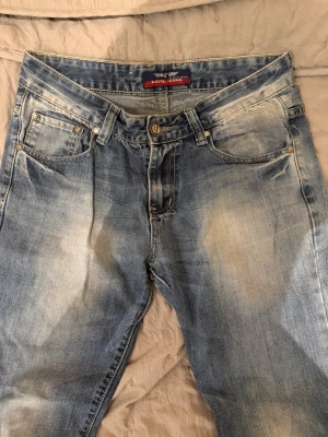 Blå jeans från Navil Jeans W30 L32 - Snygga ljusblå jeans från Navil Jeans med coola slitningar och kontrastsömmar. Modellen har fem fickor, bälteshällor och detaljerade bakfickor med röd och vit söm. Passar dig som gillar en avslappnad och modern stil.