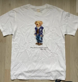 Vit Polo Bear t-shirt från Ralph Lauren - Vit t-shirt från Polo Ralph Lauren med ikoniskt Polo Bear-tryck på bröstet. Klassisk rund hals och korta ärmar. Tillverkad i mjuk bomull, perfekt för en chill och stilren look. Snygg detalj med björnen i collegejacka och jeans.