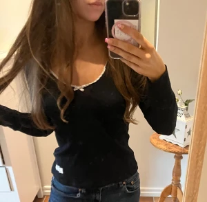 Svart ribbad långärmad tröja Odd Molly - Supersöt svart ribbad långärmad tröja från Odd Molly med vit spetskant och liten rosett vid v-ringningen. På ena ärmen finns en broderad fjäril i färgerna blå, beige och vit. Tröjan är figurnära och har mjukt bomullsmaterial, perfekt för dig som gillar detaljer.