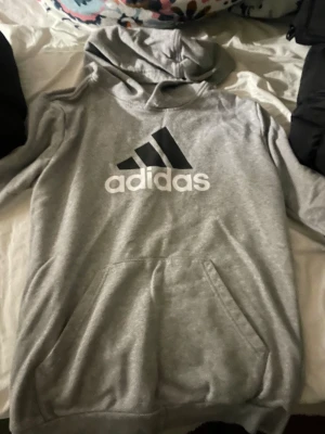 Grå Adidas hoodie med logga - Säljer en grå hoodie från Adidas med stor svart och vit logga på bröstet. Tröjan har huva, magficka och långa ärmar. Perfekt för chill eller träning, riktigt skön och klassisk stil.