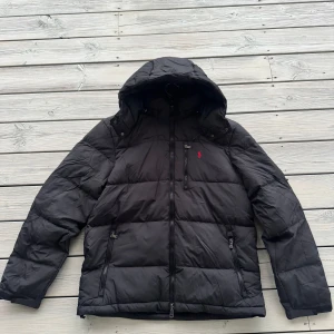 Ralph Lauren Dunjacka - Svart pufferjacka från Polo Ralph Lauren med huva, flera dragkedjefickor och ikonisk röd logga på bröstet. Jackan är quiltad och har en normal passform, perfekt för kalla dagar. Jackan är lite tillplattad eftersom den hängt tillsammans med andra jackor. Men kommer puffas upp till normalt läge vid användning.