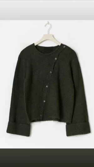 Mörkgrön stickad kofta med knappar - Mysig mörkgrön stickad kofta med rund hals och unika knappar som går snett över framsidan. 