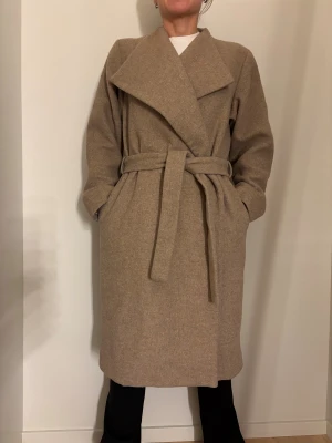 Beige Wera oversized  ullkappa  med bälte - Säljer en beige oversized kappa i omlottmodell med bred krage och knytskärp i midjan. Jackan är lång, har raka ärmar och är tillverkad i ett mjukt ullmaterial. Perfekt för dig som gillar stilrena och minimalistiska plagg. Inköpt på Åhléns för ca 1900kr sparsamt använd. Från djur- och rökfritt hem. Jag är 166cm lång och har vanligtvis 36 så denna lite oversized.