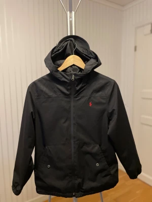 Svart vindjacka från Polo Ralph Lauren - Svart vindjacka från Polo Ralph Lauren med huva och dragkedja framtill. Jackan har två fickor med knappstängning och den klassiska röda Polo-loggan på bröstet. Tillverkad i polyester och har elastiska muddar vid ärmsluten. Perfekt för blåsiga dagar.