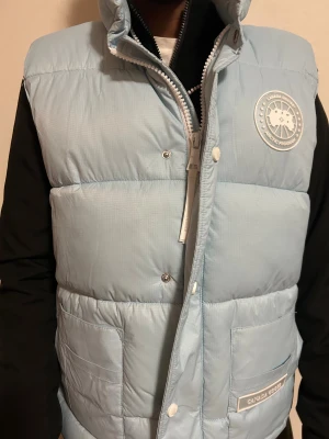 Ljusblå dunväst från Canada Goose, M - Snygg ljusblå dunväst från Canada Goose med hög krage, två stora fickor framtill och vit logotyp på bröstet. Västen har quiltad design och stängs med dragkedja och tryckknappar. Perfekt för kalla dagar när du vill hålla stilen.