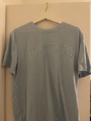 Ljusblå Brooklyn NYC t-shirt Manfinity - Snygg ljusblå t-shirt från Manfinity i storlek M med upphöjd BROOKLYN NYC-text på bröstet. Klassisk rund hals och korta ärmar. Perfekt för dig som gillar streetstyle och enkel design. Materialet är mjuk bomull som känns skönt mot huden.
