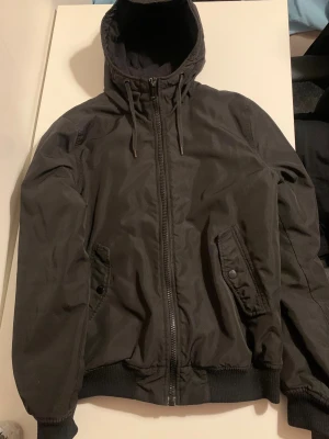 Svart bomberjacka med huva - Svart bomberjacka från DB Supply med huva och snörning. Jackan har ribbade muddar, dragkedja framtill och två fickor med knappstängning. Insidan är fodrad med glansigt material som ger extra komfort. Perfekt för dig som gillar stilren och enkel look.