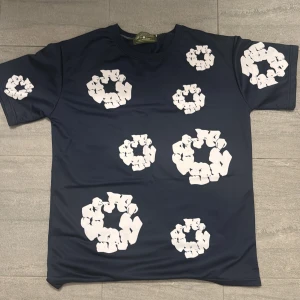 Mörkblå t-shirt från Denim Tears - Snygg mörkblå t-shirt från Denim Tears med unika vita blommönster tryckta över hela plagget. Klassisk passform med rund hals och korta ärmar. Perfekt statement piece för dig som vill sticka ut med stil.