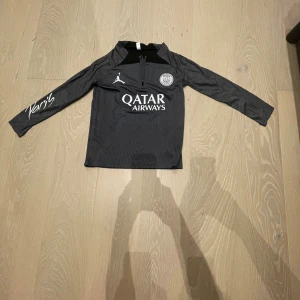 PSG långärmad träningströja Jordan - Mörkgrå långärmad fotbollströja från PSG och Jordan med halv dragkedja, Paris Saint-Germain-logga på bröstet och ärmen, samt Qatar Airways-tryck framtill. Tillverkad i lätt och sportigt material, perfekt för träning. Snygga detaljer och modern passform. Storlek runt 13-140
