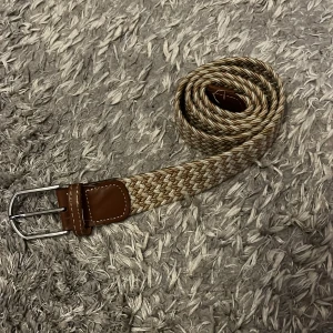 Beige och brun flätat bälte - Snyggt flätat bälte i beige och bruna toner med detaljer i brunt skinn och silverspänne. Perfekt accessoar för att lyfta din outfit med en stilren och klassisk look. Bältet har en flexibel design tack vare det flätade materialet.
