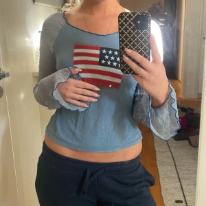 Blå croppad topp med USA-flagga - Köpt i NewYork. Säljer en blå croppad topp med långärmade, transparenta ärmar och vågiga kanter. Framsidan har ett tryck av den amerikanska flaggan. 