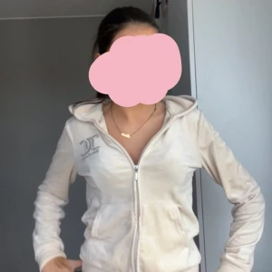 Juicy Couture beige velour hoodie - Juicy Couture hoodie i beige velour med dragkedja framtill, huva och broderad logga på bröstet samt stor brodyr på ryggen. Mjuk och skön med fickor på sidorna och klassisk Juicy-stil. Perfekt för dig som gillar comfy och trendigt.