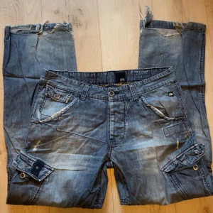 Blå cargopants - Säljer ett par blå cargopants från Jack & Jones med flera fickor, knappar och coola detaljer. Byxorna har bred passform och är tillverkade i jeansmaterial. Slitna vid änden på benen men inget man tänker på. Jag är ~170cm och dem passar bra. Skriv om ni har frågor💕