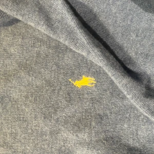 Blå stickad tröja från Polo Ralph Lauren - Snygg blå stickad tröja från Polo Ralph Lauren i storlek XL. Tröjan har rund halsringning, ribbade muddar och en gul broderad logga på bröstet. Perfekt för dig som gillar klassisk och stilren design. Använd ett fåtal gånger