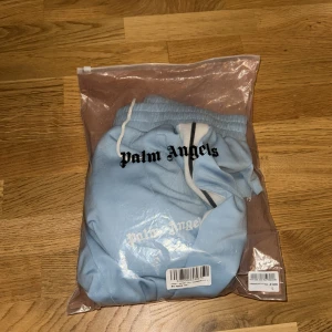 Palm Angels ljusblå trackpants S - Palm Angels ljusblå trackpants med vita och svarta ränder längs sidorna. Byxorna har elastisk midja med vit snörning och Palm Angels-logga tryckt på vänster ben. Tillverkade i ett mjukt, syntetiskt material som ger en sportig vibe.