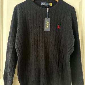 Svart kabelstickad tröja Polo Ralph Lauren - Kabelstickad svart tröja från Polo Ralph Lauren med klassisk rund halsringning och röd broderad logga på bröstet. Långärmad modell i mjuk ull, perfekt för kyliga dagar. Snygg och tidlös design som funkar till allt.