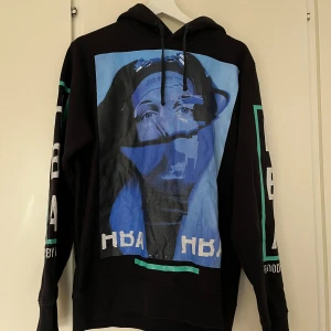 Svart hoodie från Hood By Air med tryck - Svart hoodie från Hood By Air med stort grafiskt print framtill och textdetaljer på ärmarna. Tröjan har huva med snörning och ribbade muddar. Oversized vibe. Trycket är något urtvättat på ärmarna.