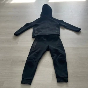 Svart Nike tech fleece  - Svart hoodiejacka från Nike med hel dragkedja framtill och två fickor med dragkedja. Jackan har en sportig look, huva och diskret Nike-logga på bröstet. Materialet är mjukt och passar perfekt för chill eller träning.