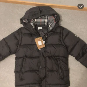 Svart pufferjacka från Burberry M - Säljer en svart pufferjacka från Burberry i storlek M. Jackan har avtagbara ärmar så den kan bäras som väst, huva med rutigt foder i grått, vitt och rött samt dragkedja och knappar framtill. Två fickor med dragkedja och klassisk Burberry-tag medföljer.