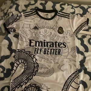 Real Madrid drakmönstrad fotbollströja - Snygg Real Madrid fotbollströja från Adidas i vitt med svart drakmönster över hela tröjan. Tröjan har klubbmärke på bröstet, svarta detaljer vid krage och ärmslut samt tryck från Emirates. Tillverkad i lätt och ventilerande material. Tröjan sitter ganska så slim. Så om du har S eller M passar den dig mycket bättre. Priset är inte hugget i sten, tveka inte med funderingar, frågor eller bud🙂