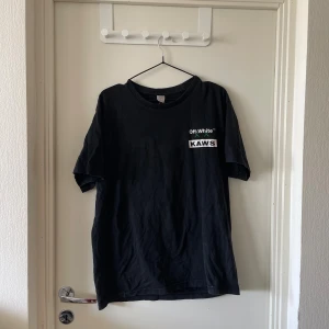 Off-White x KAWS svart t-shirt - Svart t-shirt från Off-White x KAWS med tryck på bröstet och stor grön KAWS-figur på ryggen. Klassisk passform, rund hals och coola vita detaljer. Perfekt för dig som gillar streetwear och unika samarbeten.