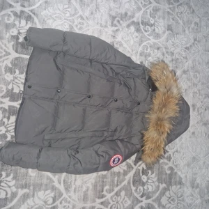 Canada Goose wyndham - Säljer en grå dunjacka från Canada Goose med ikonisk patch på ärmen. Jackan har en stor huva med fluffig pälsdetalj, knappar framtill och klassisk puffer-design. Perfekt för kalla dagar och riktigt snygg passform.