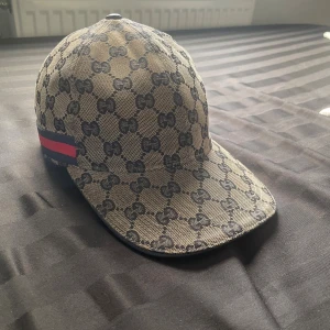 Gucci keps med GG-mönster, S - Snygg beige och svart keps från Gucci med klassiskt GG-mönster över hela kepsen. Har en justerbar rem i svart skinn baktill och en röd-blå detalj på sidan. Materialet är bomull och skinn, perfekt för dig som vill ha en lyxig streetstyle-accessoar. Pris ej hugget i sten!