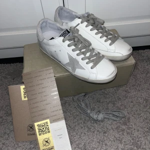 Golden Goose vita sneakers med grå stjärna - Golden Goose sneakers i vitt skinn med grå mockadetaljer och en ikonisk grå stjärna på sidan. Skorna har grå snörning, rund tå och platt sula. Märkeslogga på plösen och insidan. Snygg och clean design som passar till jeans och hoodie.