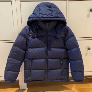 Mörkblå pufferjacka från Polo Ralph Lauren - Säljer en mörkblå pufferjacka från Polo Ralph Lauren med huva och dragkedja framtill. Jackan har två fickor med dragkedja och den klassiska röda Polo-loggan på bröstet. Första bilden från förra ägaren men har inte använts så mycket av mig heller så ingen skillnad från då och nu💕