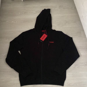 Hugo kofta - Svart kofta från HUGO med röd logga på bröstet. Tröjan har huva med snörning, hel dragkedja och långa ärmar. Perfekt för en chill och stilren look. Tillverkad i mjukt material för extra komfort.