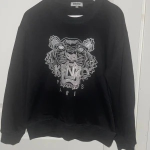Svart Kenzo sweatshirt med tiger - Cool svart sweatshirt från Kenzo med ikoniskt broderat tigerhuvud och Kenzo Paris-text i silver på bröstet. Tröjan har rund halsringning och långa ärmar. Perfekt för dig som vill sticka ut med en streetstyle-look.