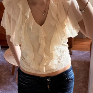 Krämvit volangblus från Kappahl XS - Säljer en krämvit blus från Kappahl i storlek XS. Blusen har v-ringning, korta ärmar och flera lager med volanger framtill och på axlarna. Materialet är tunt och lätt, perfekt för vår och sommar. Snygg att matcha med jeans eller kjol för en trendig look.