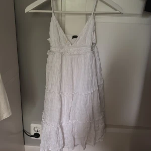Vit omlottklänning från Bershka XS - Supersöt vit kortklänning från Bershka i storlek XS. Klänningen har omlott-design med v-ringning, tunna axelband och flera volanger som ger en flowy känsla. Materialet är lätt och krinklat, perfekt för varma dagar. Justerbara axelband för bästa passform. OBS! Har en liten fläck vid uringningen, se sista bild!