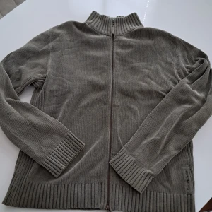 Grå (mörkt grön) kofta från Marlboro Classics - Säljer en grå ribbstickad kofta från Marlboro Classics i storlek L. Koftan har hel dragkedja framtill, hög krage och långa ärmar. Tillverkad i mjukt bomullsmaterial och har diskret logga på sidan. Perfekt för lager-på-lager under kyliga dagar.