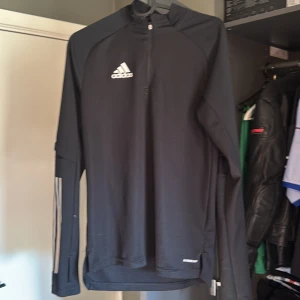 Svart Adidas långärmad träningströja - Svart långärmad träningströja från Adidas med vitt logotryck på bröstet och diskreta ränder på ena ärmen. Tröjan har half zip och är tillverkad i ett lätt syntetmaterial som andas, perfekt för träning eller sport.