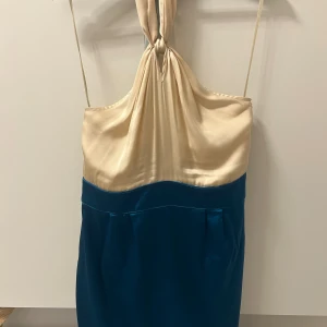 Tvåfärgad halterneckklänning från Oasis - Supersnygg halterneckklänning från Belle by Oasis i glansigt material. Överdelen är champagnefärgad och underdelen är blå, med en elegant knut i nacken och smala axelband. Klänningen har dragkedja i sidan och en figurnära passform som ger en chic siluett.