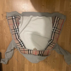 Grå Burberry hoodie med dragkedja - Säljer en grå hoodie från Burberry med klassiskt rutigt foder i huvan. Tröjan har dragkedja framtill, snörning i huvan och två fickor. Perfekt för dig som vill ha en stilren och lyxig look med ikoniska Burberry-detaljer.