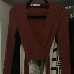 Rostbrun omlottopp från Zara - Säljer en ribbad omlottopp från Zara i en snygg rostbrun färg. Toppen har långa ärmar, croppad passform och knytband i midjan. Djup v-ringning och stretchigt material gör den både trendig och bekväm. Perfekt att styla med höga jeans eller kjol.