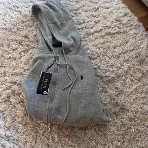 Grå hoodie från Polo Ralph Lauren - Snygg grå hoodie från Polo Ralph Lauren med dragkedja och huva. Klassisk logga broderad på bröstet och praktiska fickor framtill. Mjuk bomullsblandning och justerbar snörning i huvan. Perfekt för chill dagar eller när du vill ha en clean look.