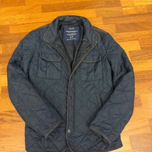 Quiltad mörkblå jacka American Eagle - Snygg quiltad mörkblå jacka från American Eagle Outfitters. Jackan har två bröstfickor med lock, två sidofickor, dragkedja och knappar framtill. Perfekt för dig som gillar stilren och klassisk look.